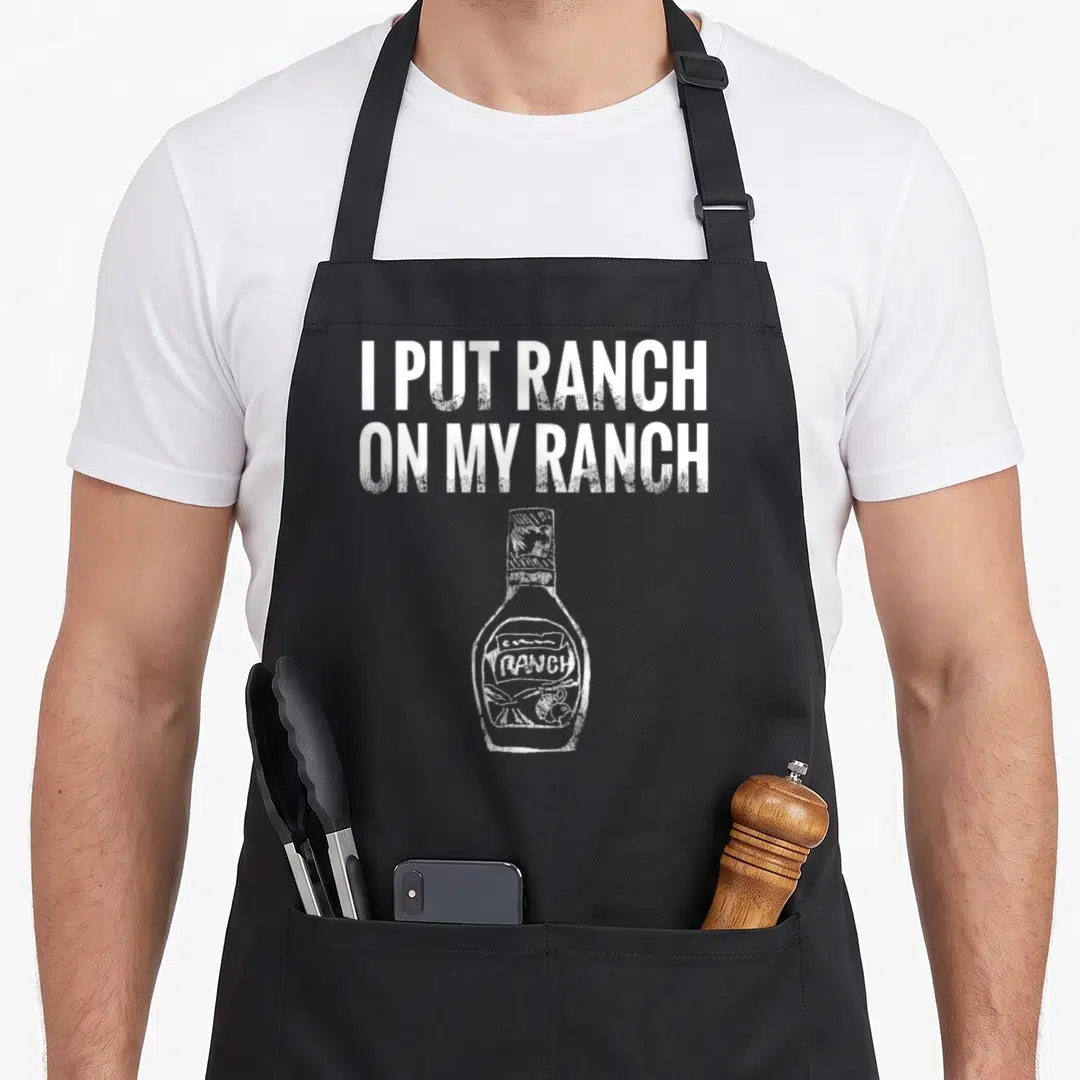 Funny ranch vibes apron - Distressed white text I PUT RANCH ON MY RANCH above a hand drawn ranch dressing bottle sketch — shown on a black cotton canvas apron against a white background
