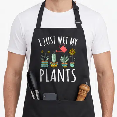 Plant wet gardener apron - I JUST WET MY PLANTS text above four potted plants and red watering can — shown on a black cotton canvas apron against a white background