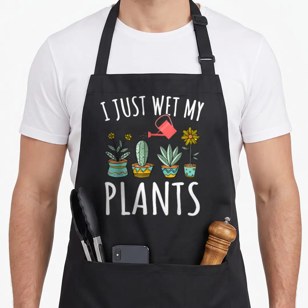 Plant wet gardener apron - I JUST WET MY PLANTS text above four potted plants and red watering can — shown on a black cotton canvas apron against a white background