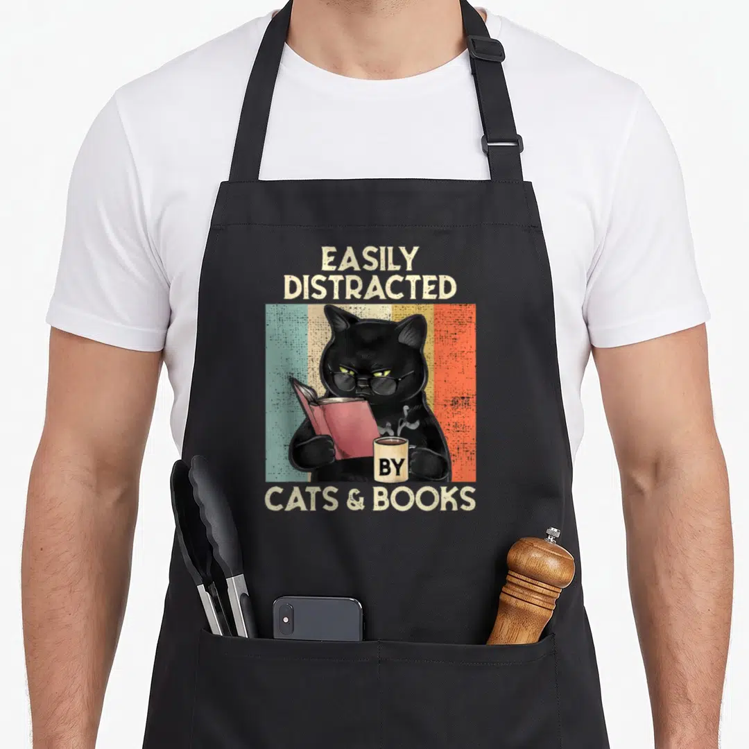 Distracted book cat apron - Black cat with glasses holding red book and mug with retro stripes and text — shown on a black cotton canvas apron against a white background