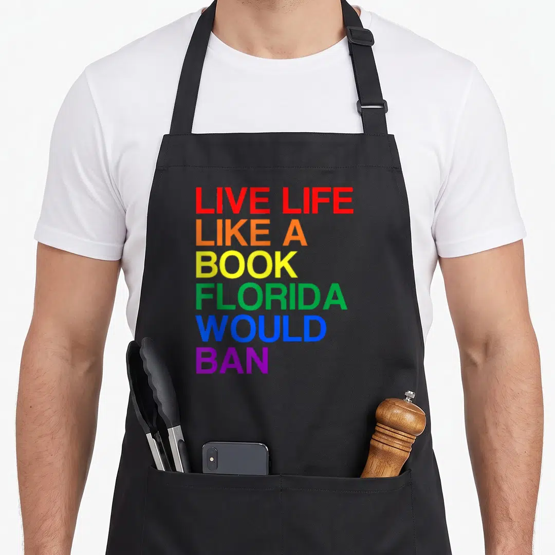 Live rainbow apron - Stacked rainbow text reading Live Life Like A Book Florida Would Ban — shown on a black cotton canvas apron against a white background