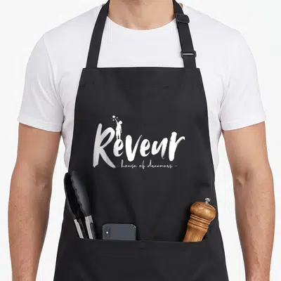 Funny dreamer teacher apron - White Rêveur text with child reaching for stars and house of dreamers subtitle on black — shown on a black cotton canvas apron against a white background