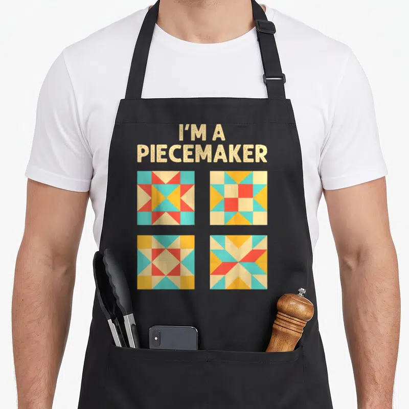 Quilting knitting apron - I'M A PIECEMAKER text above four beige red yellow and blue quilt squares in a grid — shown on a black cotton canvas apron against a white background