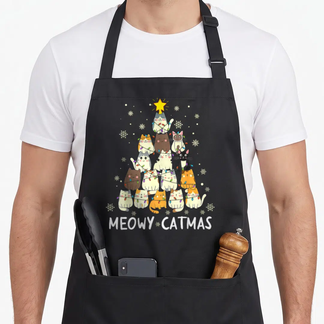 Meowy christmas tree apron - Fourteen cats stacked in pyramid shape with MEOWY CATMAS text and yellow star — shown on a black cotton canvas apron against a white background
