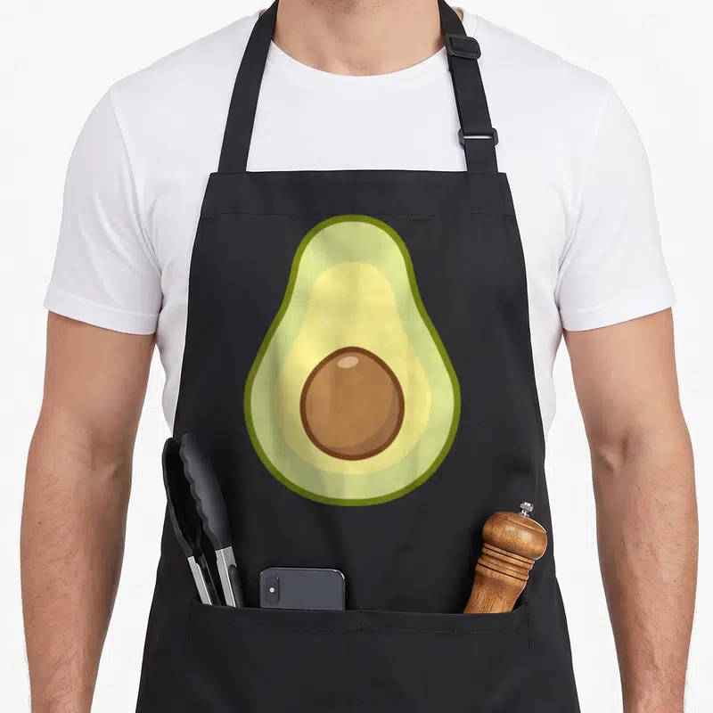 Avocado vegan apron - Cartoon sliced avocado facing forward with green yellow brown pit — shown on a black cotton canvas apron against a white background