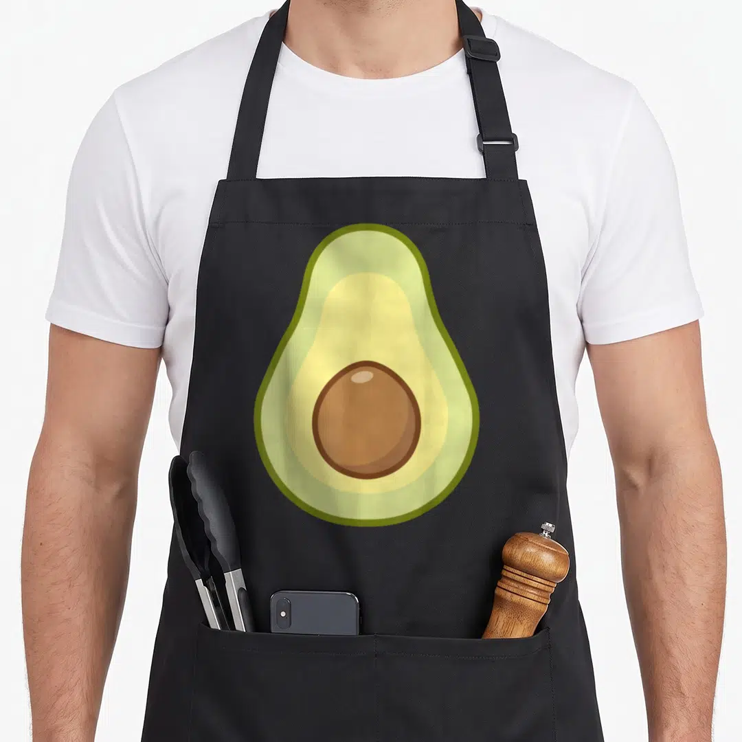 Avocado vegan apron - Cartoon sliced avocado facing forward with green yellow brown pit — shown on a black cotton canvas apron against a white background