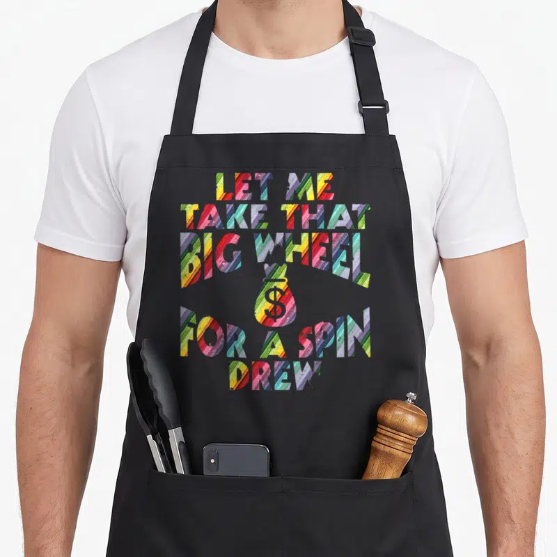 Spin wheel apron - Multicolored striped text reading LET ME TAKE THAT BIG WHEEL FOR A SPIN DREW with central money bag — shown on a black cotton canvas apron against a white background