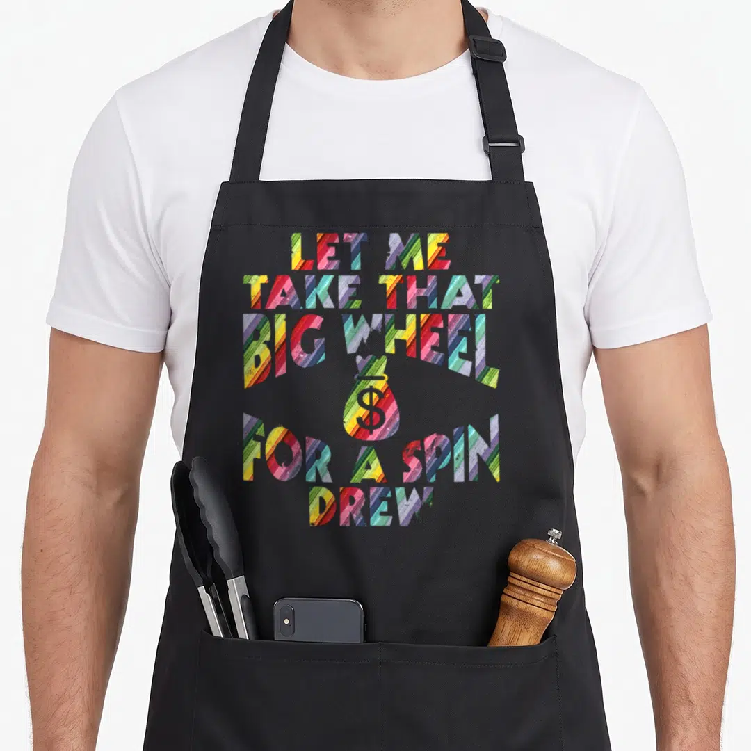 Spin wheel apron - Multicolored striped text reading LET ME TAKE THAT BIG WHEEL FOR A SPIN DREW with central money bag — shown on a black cotton canvas apron against a white background