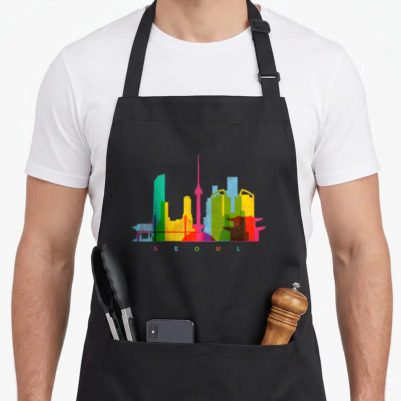 Seoul colourful apron - Colorful Seoul skyline with matching letters S E O U L in teal green yellow pink red blue — shown on a black cotton canvas apron against a white background