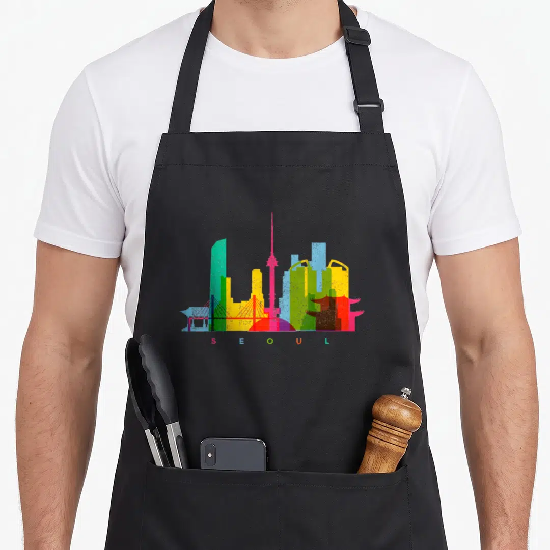 Seoul colourful apron - Colorful Seoul skyline with matching letters S E O U L in teal green yellow pink red blue — shown on a black cotton canvas apron against a white background