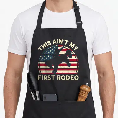 Cowboy american flag apron - THIS AIN'T MY FIRST RODEO text over circular flag and cowboy silhouette in beige and grey — shown on a black cotton canvas apron against a white background