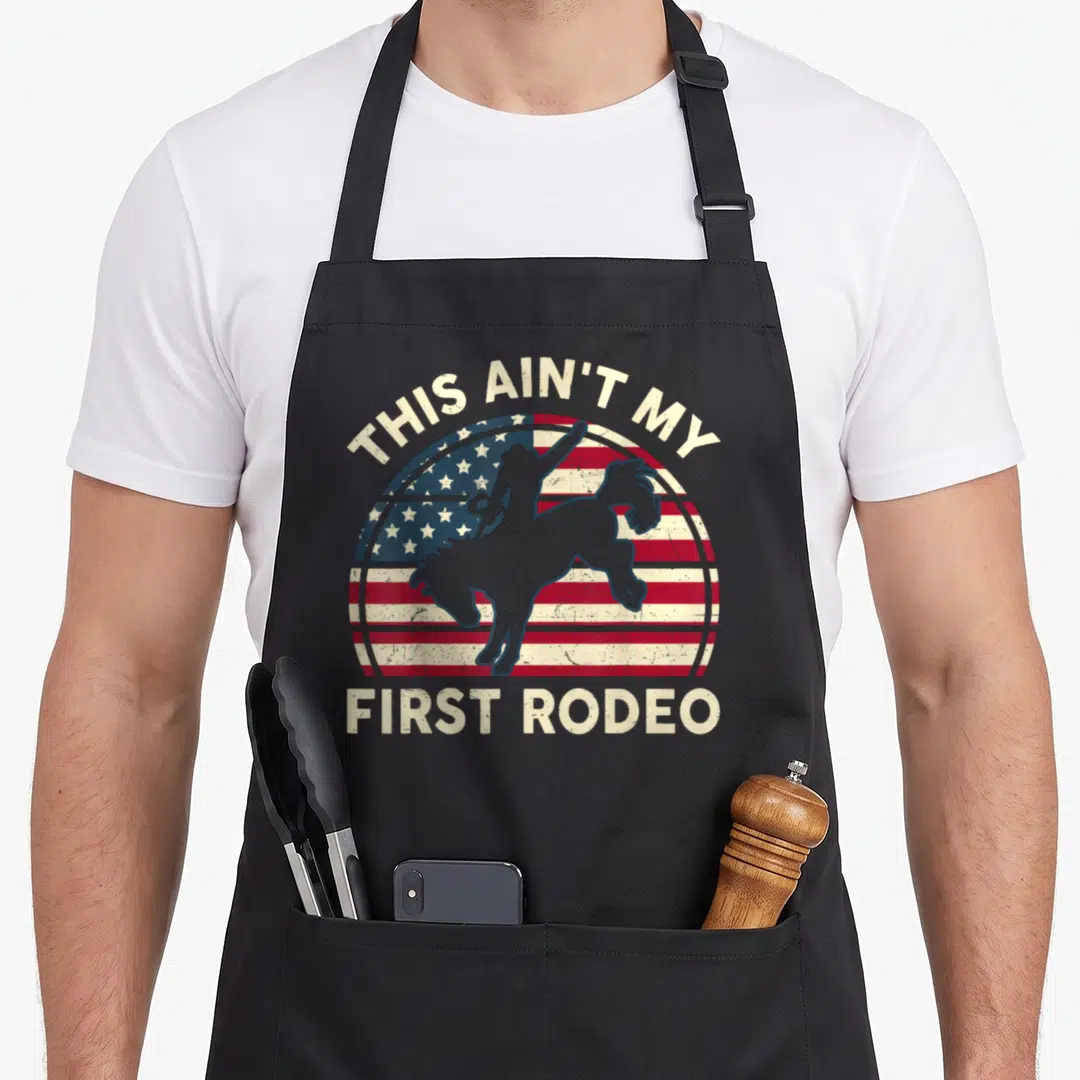 Cowboy american flag apron - THIS AIN'T MY FIRST RODEO text over circular flag and cowboy silhouette in beige and grey — shown on a black cotton canvas apron against a white background