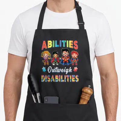 Ability teacher art apron - ABILITIES Outweigh DISABILITIES text with four superhero kids and puzzle pieces in rainbow — shown on a black cotton canvas apron against a white background