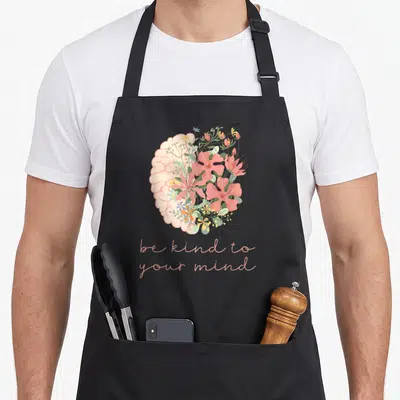 Kind anxiety awareness apron - Brain with pink floral right half and peach text be kind to your mind — shown on a black cotton canvas apron against a white background