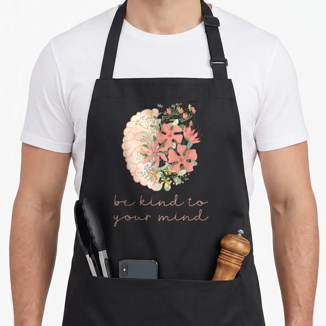 Kind anxiety awareness apron - Brain with pink floral right half and peach text be kind to your mind — shown on a black cotton canvas apron against a white background