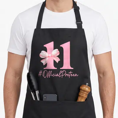 Coquette girly apron - Pink number 11 with light pink bow and #OfficialPreteen text — shown on a black cotton canvas apron against a white background