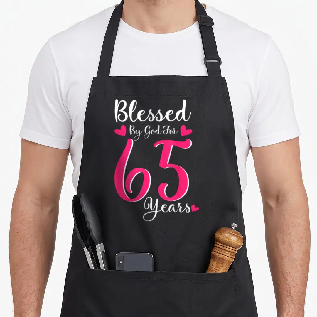 Tea birthday apron - White and pink text reading Blessed By God For 65 Years with three heart icons — shown on a black cotton canvas apron against a white background