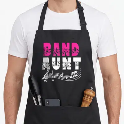 Aunt band mom apron - Stacked pink and white BAND AUNT text filled with musical notes and a flowing staff wave below — shown on a black cotton canvas apron against a white background