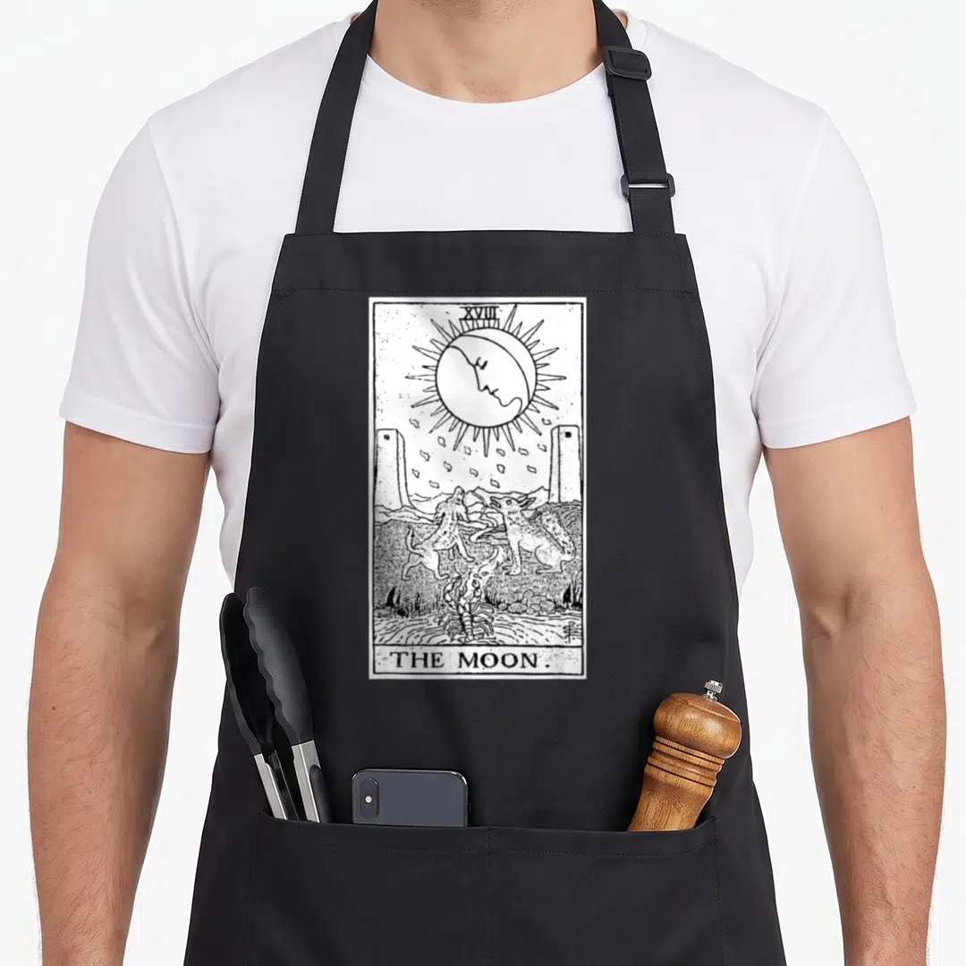 Wiccan craft apron - Black and white line art of The Moon tarot card with moon face, two animals, and crayfish — shown on a black cotton canvas apron against a white background