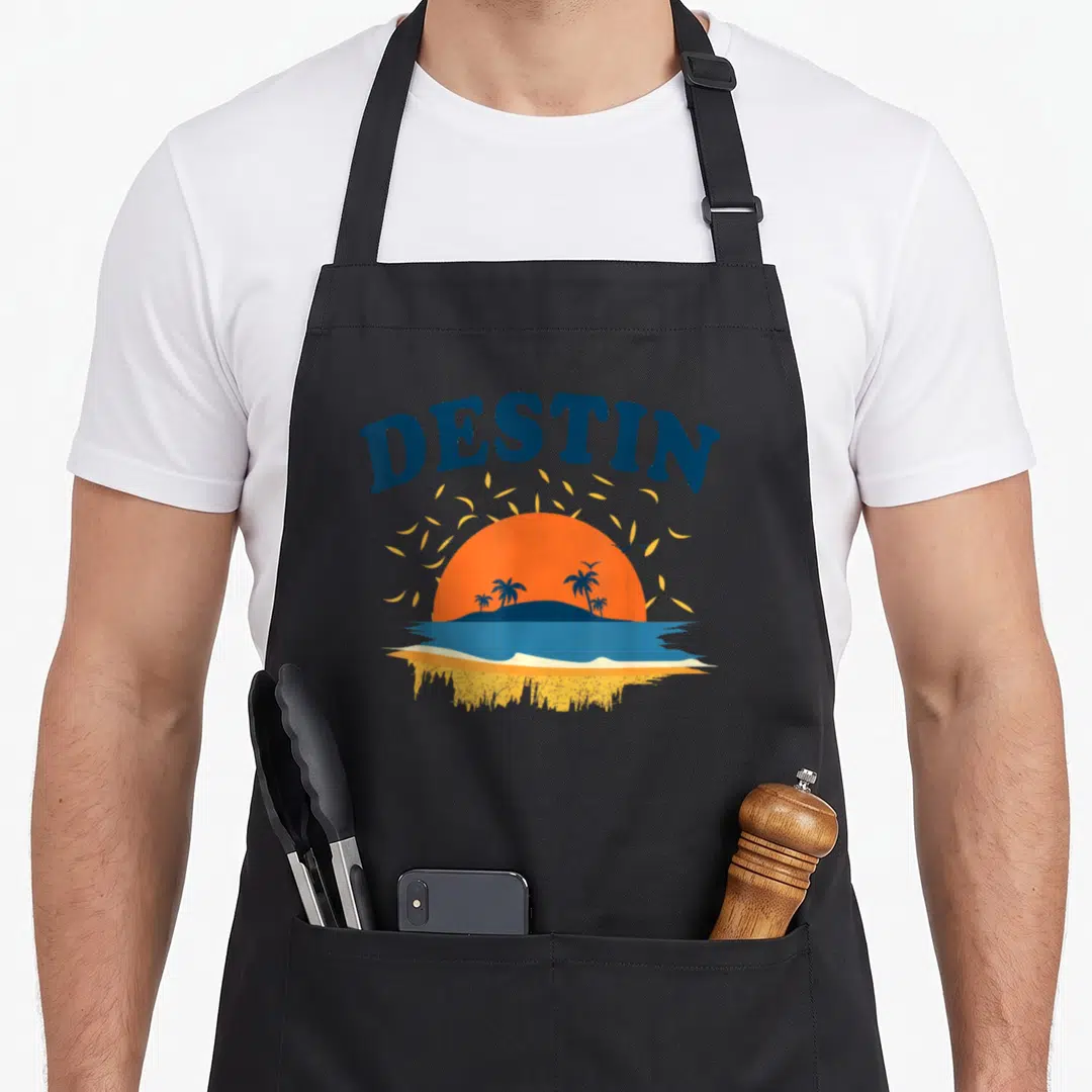 Destin tourist florida apron - Minimalist orange sunset over blue water with palm trees and arched DESTIN text — shown on a black cotton canvas apron against a white background