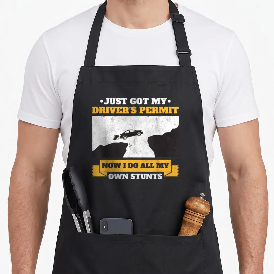 Driver congratulations apron - Car flying off cliff with Just Got My Drivers Permit text in yellow and black — shown on a black cotton canvas apron against a white background