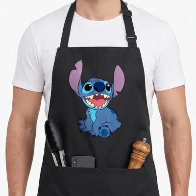 Sitting stitch apron - Blue cartoon creature sitting with purple ears and wide toothy grin — shown on a black cotton canvas apron against a white background