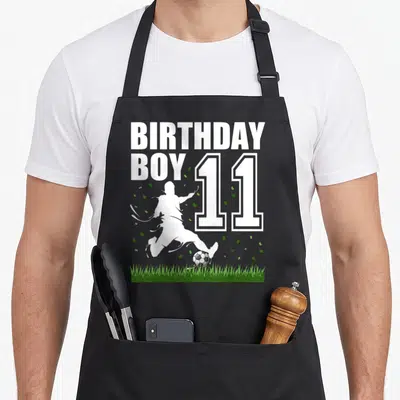 Theme 11th birthday apron - BIRTHDAY BOY 11 text with soccer player silhouette kicking ball on grass — shown on a black cotton canvas apron against a white background