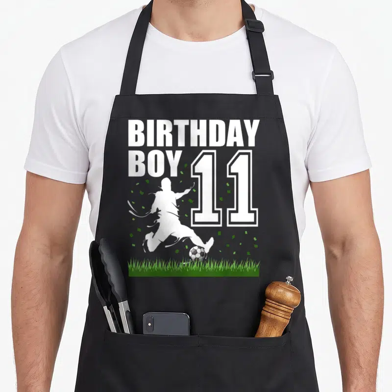 Theme 11th birthday apron - BIRTHDAY BOY 11 text with soccer player silhouette kicking ball on grass — shown on a black cotton canvas apron against a white background