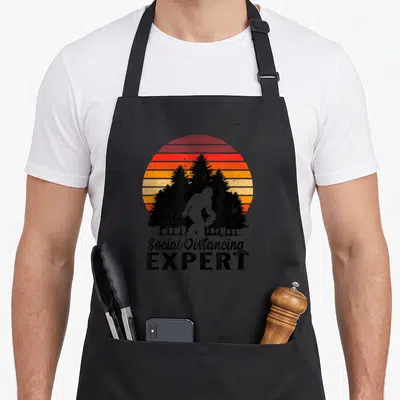 Bigfoot at home aprons - Retro Bigfoot silhouette against striped sun with Social Distancing Expert text — shown on a black cotton canvas apron against a white background