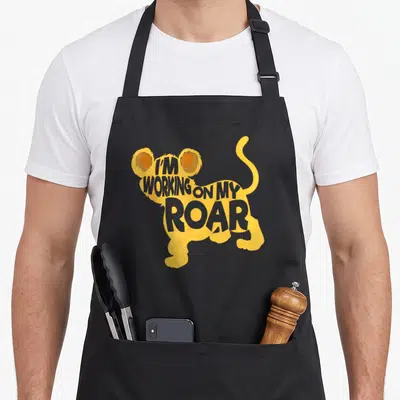 Whisperer lion apron - Yellow and orange lion silhouette containing stacked grey text reading I'M WORKING ON MY ROAR — shown on a black cotton canvas apron against a white background
