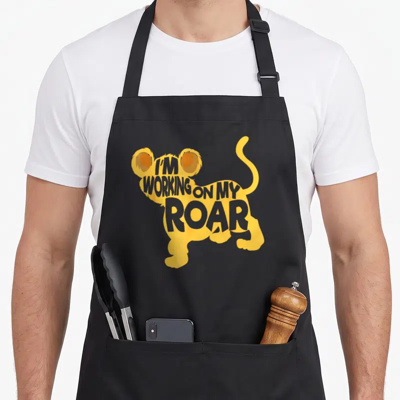 Whisperer lion apron - Yellow and orange lion silhouette containing stacked grey text reading I'M WORKING ON MY ROAR — shown on a black cotton canvas apron against a white background