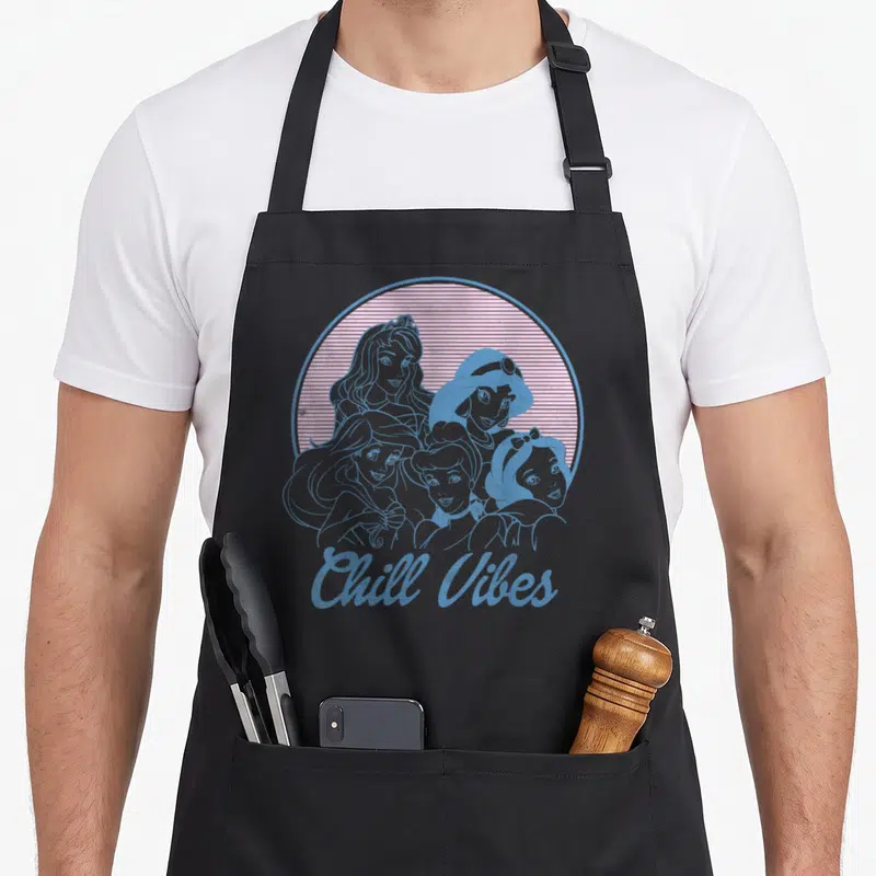Shot princess apron - Five princess silhouettes in striped circle with Chill Vibes text in blue and pink retro style — shown on a black cotton canvas apron against a white background