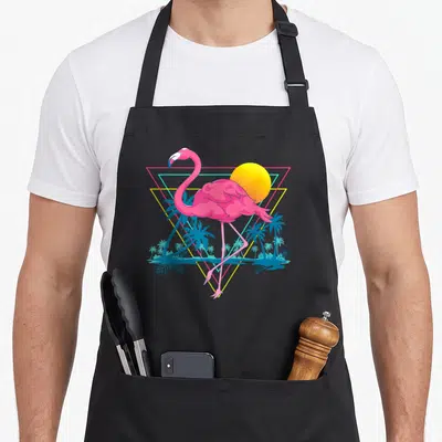 Beach pink flamingo apron - Pink flamingo standing before yellow sun and blue palms inside neon triangle — shown on a black cotton canvas apron against a white background