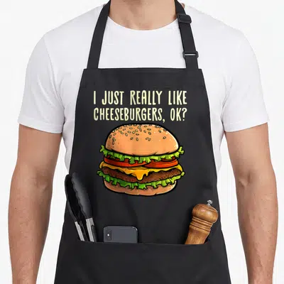 Cheeseburger burger apron - I Just Really Like Cheeseburgers, Ok? text above a cartoon cheeseburger with tan bun, green — shown on a black cotton canvas apron against a white background