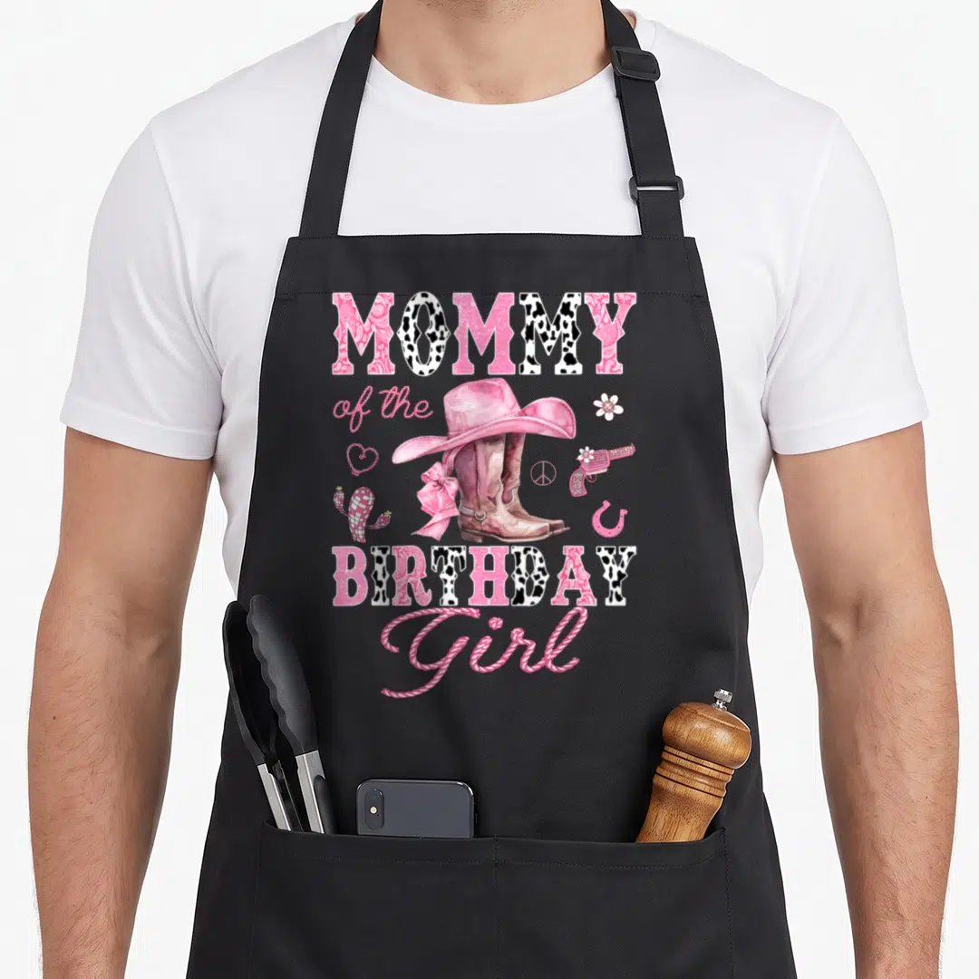 Birthday western cowgirl aprons - MOMMY of the BIRTHDAY Girl text surrounding pink cowboy boots, hat, cactus — shown on a black cotton canvas apron against a white background