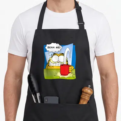 Funny bean apron - Cartoon cat holds red mug, white coffee streams up, BEAN ME! thought bubble, blue background — shown on a black cotton canvas apron against a white background