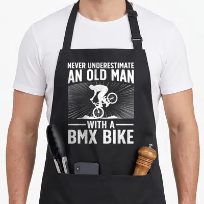 Bmx grandpa apron - White distressed text reading never underestimate an old man with a bmx bike around a stunt rider — shown on a black cotton canvas apron against a white background
