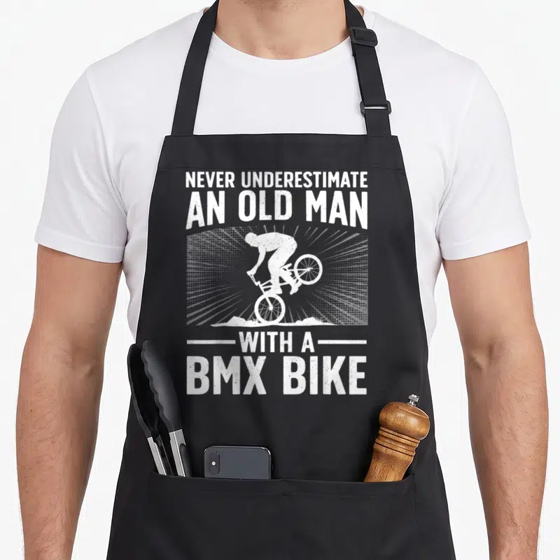 Bmx grandpa apron - White distressed text reading never underestimate an old man with a bmx bike around a stunt rider — shown on a black cotton canvas apron against a white background