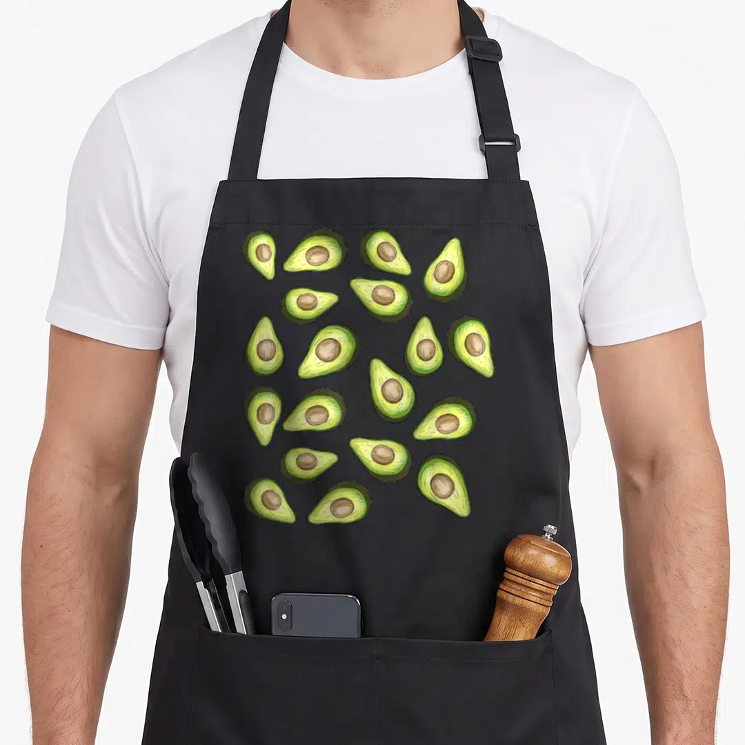 Pattern avocado apron - Fifteen cartoon avocado halves with pits in green yellow and brown scattered on black fabric — shown on a black cotton canvas apron against a white background