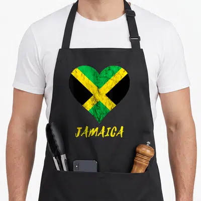 Jamaica travel aprons - Distressed heart with green yellow black flag pattern above yellow JAMAICA text — shown on a black cotton canvas apron against a white background