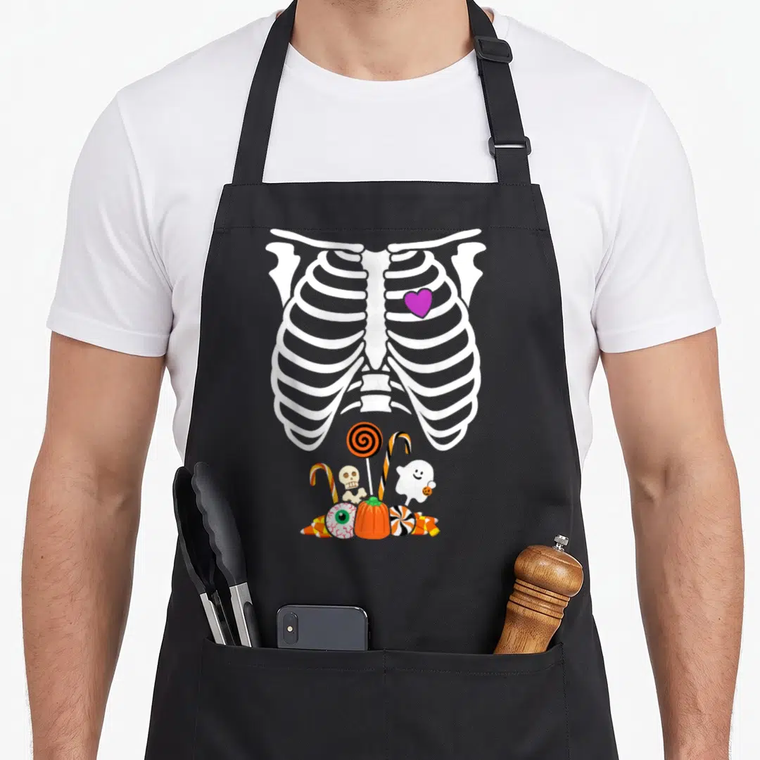 Halloween x ray apron - White ribcage with purple heart above pile of cartoon ghost pumpkin candy and eyeball — shown on a black cotton canvas apron against a white background