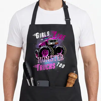 Funny monster truck aprons - Magenta monster truck graphic with white and pink text reading GIRLS LIKE MONSTER TRUCKS — shown on a black cotton canvas apron against a white background