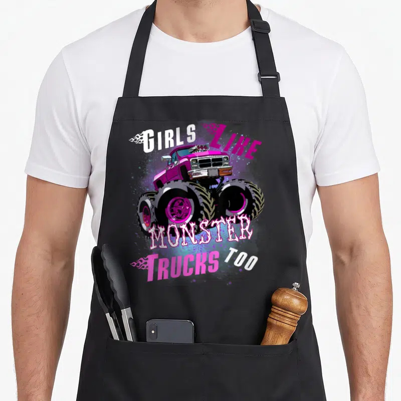 Funny monster truck aprons - Magenta monster truck graphic with white and pink text reading GIRLS LIKE MONSTER TRUCKS — shown on a black cotton canvas apron against a white background
