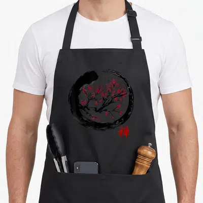 Japanese buddhist faith apron - Black brushstroke circle with red cherry blossoms and red Zen character below — shown on a black cotton canvas apron against a white background