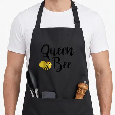 Beehive queen bee apron - Cartoon bee wearing a crown next to stacked Queen Bee text in black and yellow — shown on a black cotton canvas apron against a white background