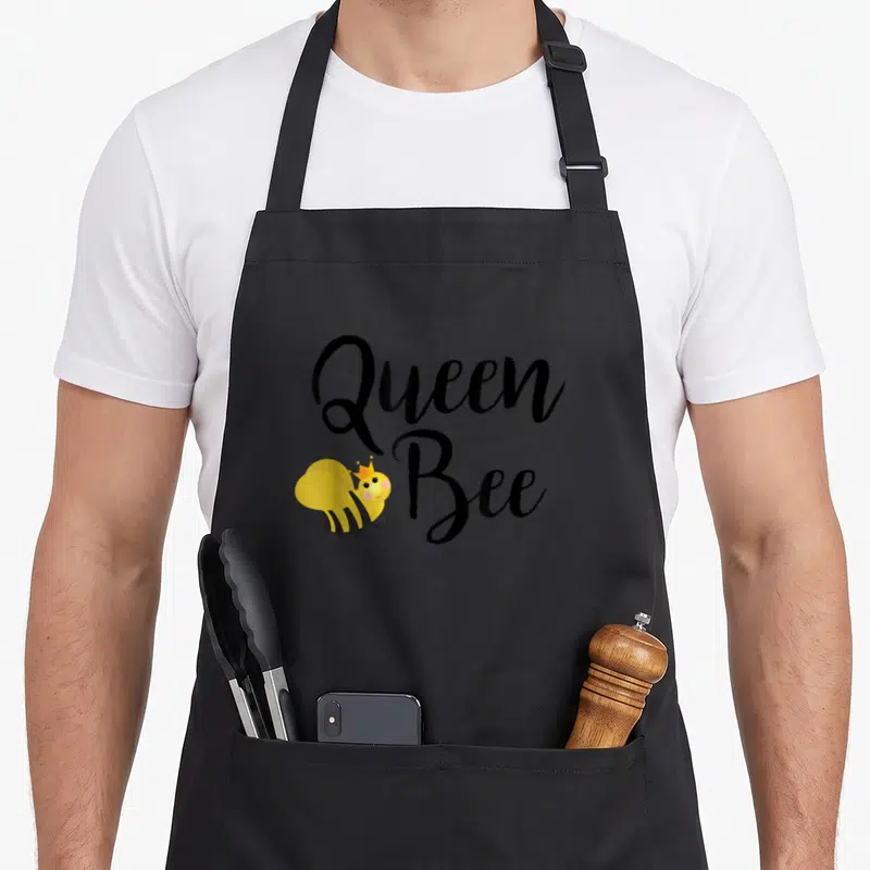 Beehive queen bee apron - Cartoon bee wearing a crown next to stacked Queen Bee text in black and yellow — shown on a black cotton canvas apron against a white background