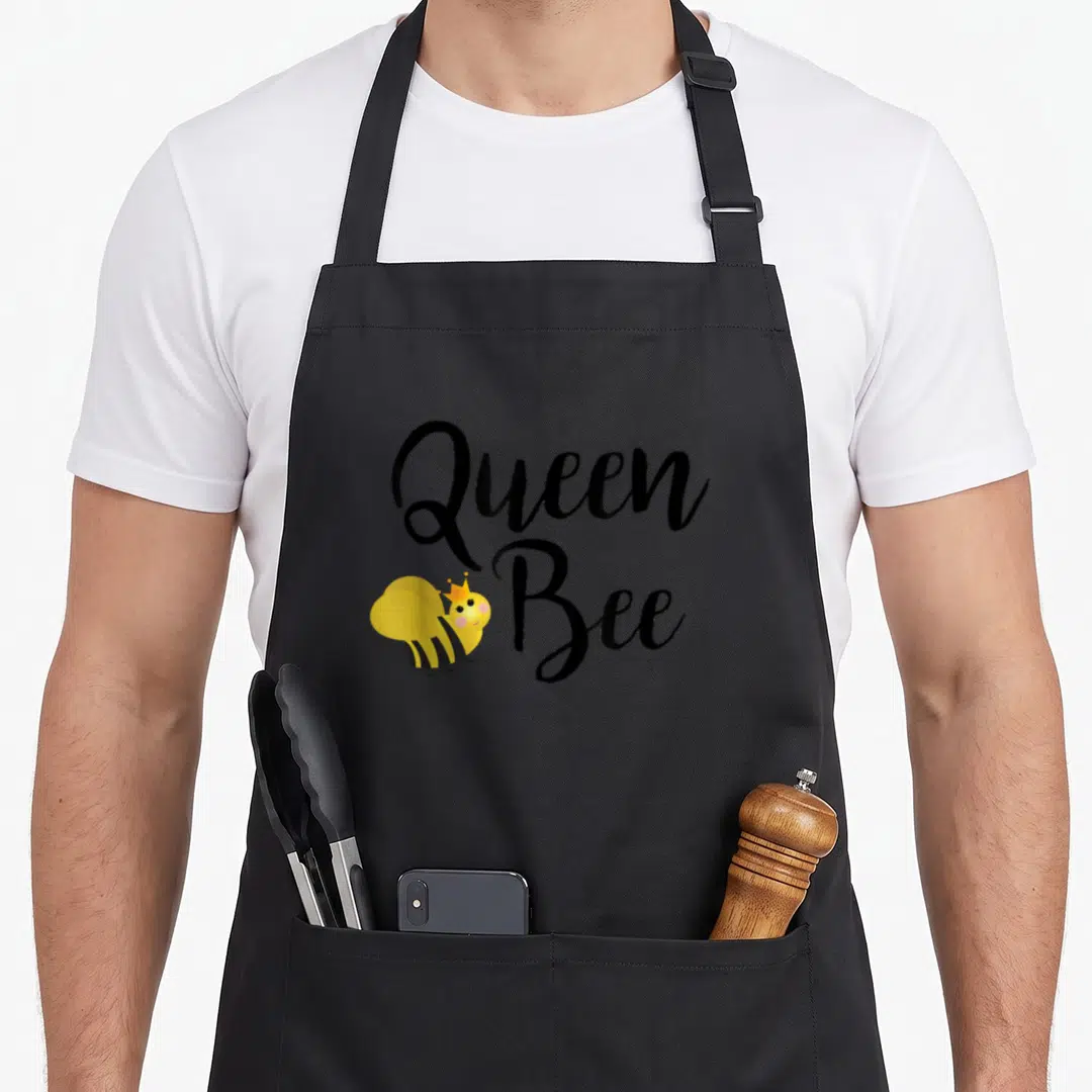 Beehive queen bee apron - Cartoon bee wearing a crown next to stacked Queen Bee text in black and yellow — shown on a black cotton canvas apron against a white background