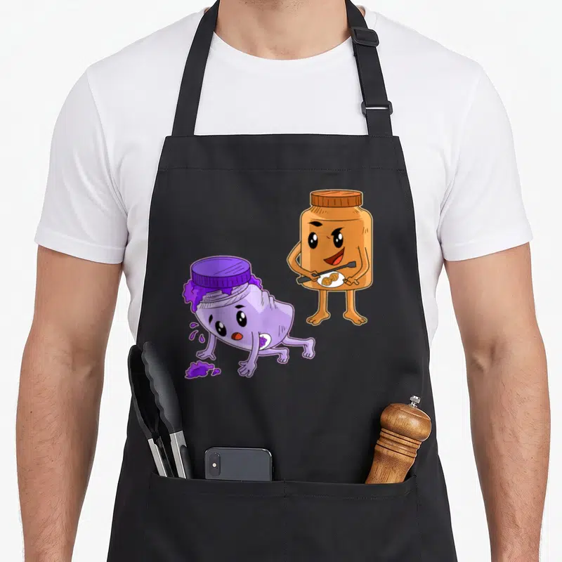 Spread butter apron - Cartoon brown peanut butter jar holding spoon next to slumped purple jam jar with spill — shown on a black cotton canvas apron against a white background