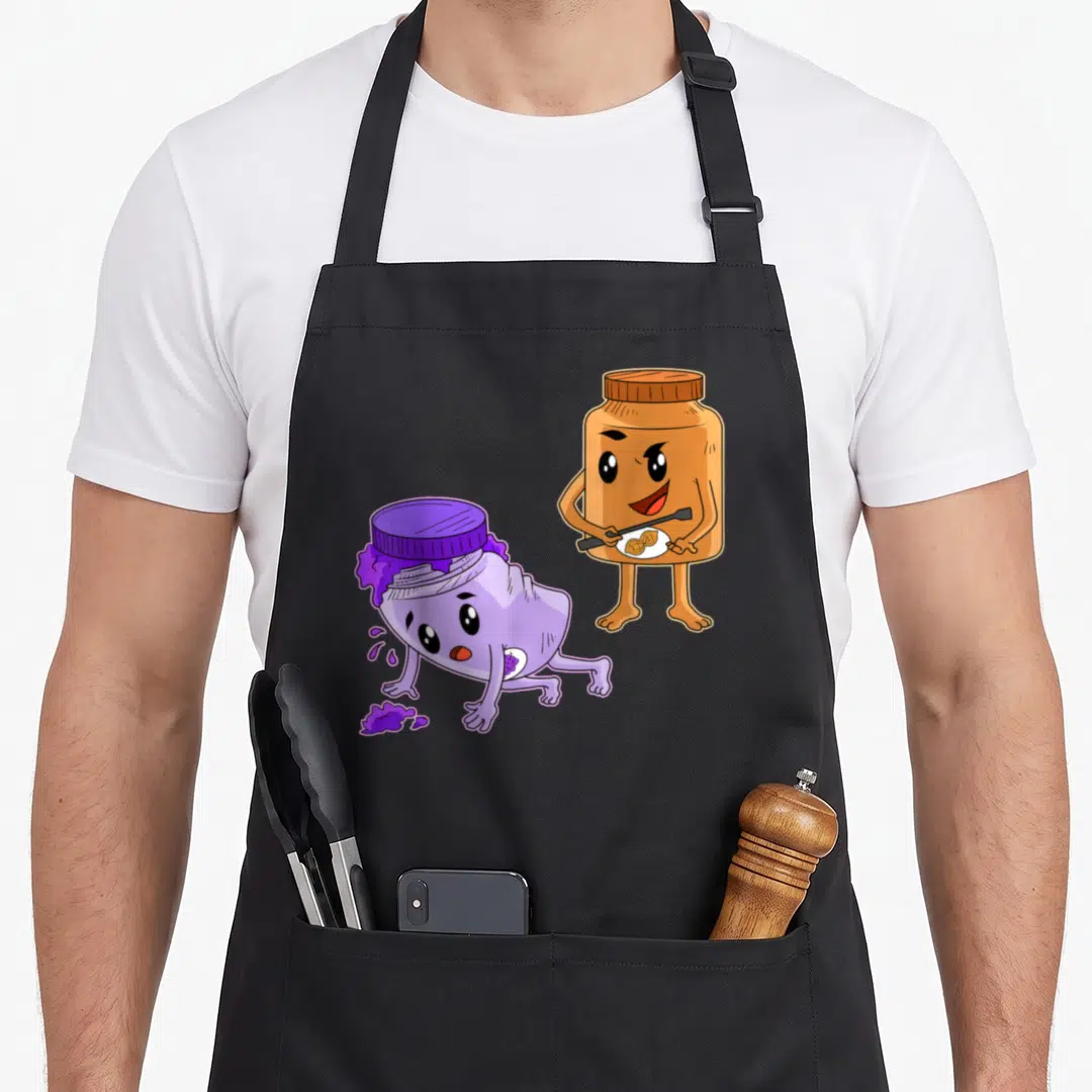 Spread butter apron - Cartoon brown peanut butter jar holding spoon next to slumped purple jam jar with spill — shown on a black cotton canvas apron against a white background