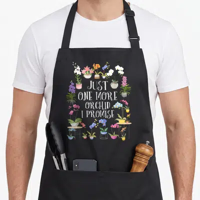 Orchid plant apron - Cartoon orchids in pots surrounding white text reading JUST ONE MORE ORCHID I PROMISE — shown on a black cotton canvas apron against a white background